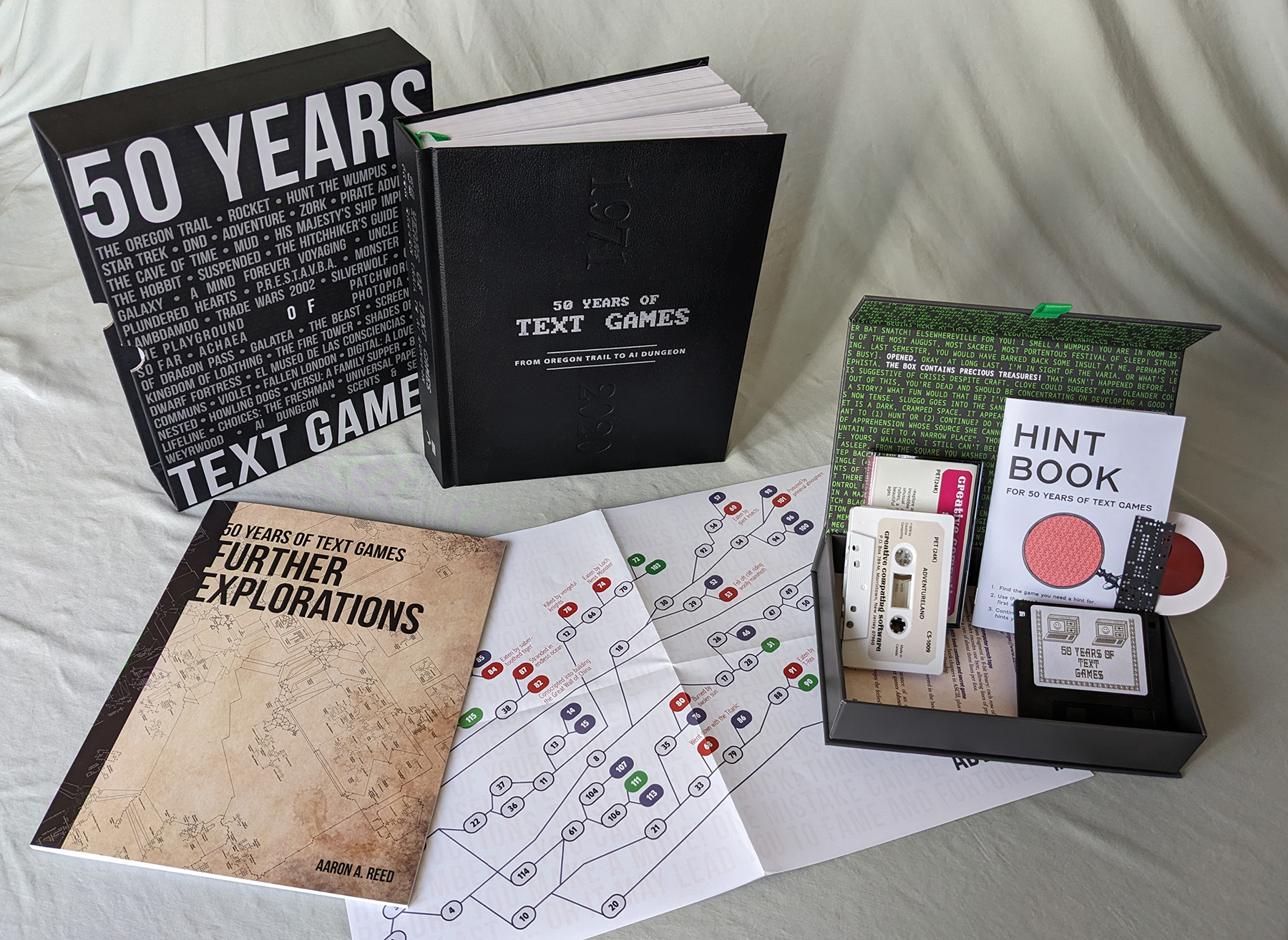 The full Ultimate Collector's Edition contents, spread out, including the slipcased hardcover edition of 50 Years of Text Games, the bonus book Further Explorations, a map of choice points in the first Choose Your Own Adventure book, and the feelies box, containing a floppy disk, a hint book with secret decoder wand, a replica of the 1978 cassette tape release of Adventureland and Pirate Adventure, and a strip of 1970s computer punch tape.